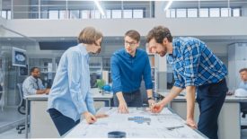 In the industrial engineering facility, a female designer works with the industrial engineer and master technician, with two people working at their desks in the background.