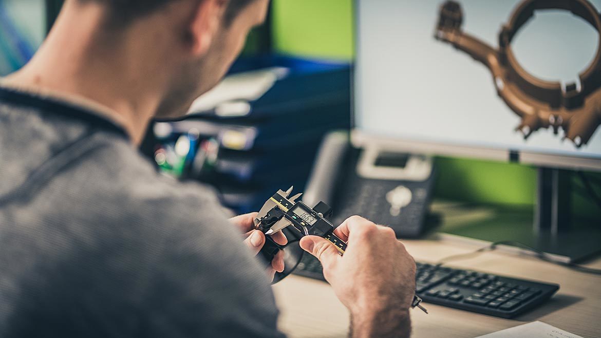 Over-the-shoulder view of an engineer measuring a machine part while working on a 3D model of it.