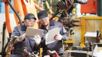 Two engineers in a factory having a discussion and looking at a tablet and clipboard.