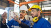 Engineer wearing safety helmet and glasses, explaining the project diagram using a futuristic tablet, with a manager in the manufacturing factory.