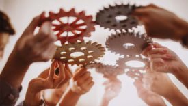 People's hands joining different metal cog wheel together.