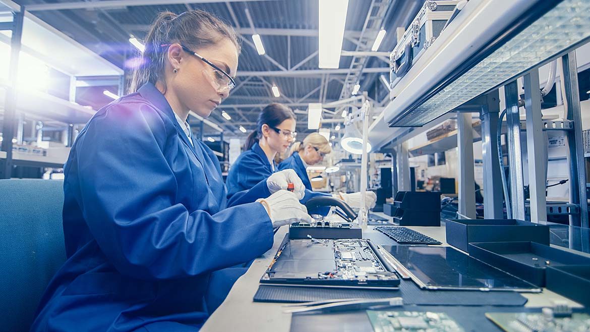 Women electronics factory workers in blue work coat and protective glasses are assembling laptop motherboards with a screwdriver.