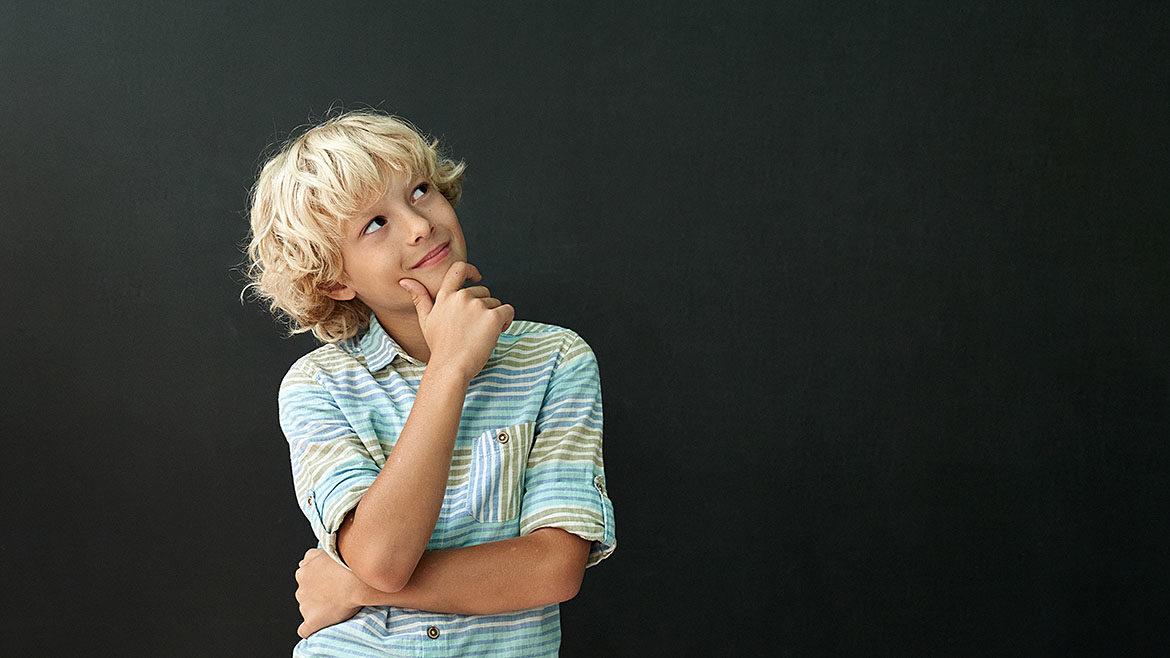 A blonde-haired boy in a striped shirt is looking up, smiling, with his right hand on his chin, with a black background.