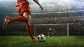 A male soccer player kicks a ball, and a soccer goalie jumps in motion for a ball while defending his gates in a wide-angle panoramic image of an outdoor soccer stadium or arena full of spectators under a sunny sky.