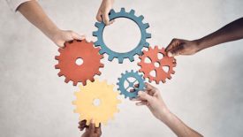 Group of five businesspeople holding and fitting gears together in an office at work. United business professionals having fun connecting gear pieces during a meeting