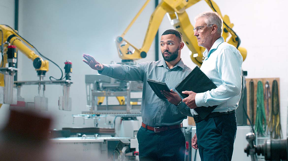 Robotics and planning with two engineers collaborating in a warehouse with robotics in the background.