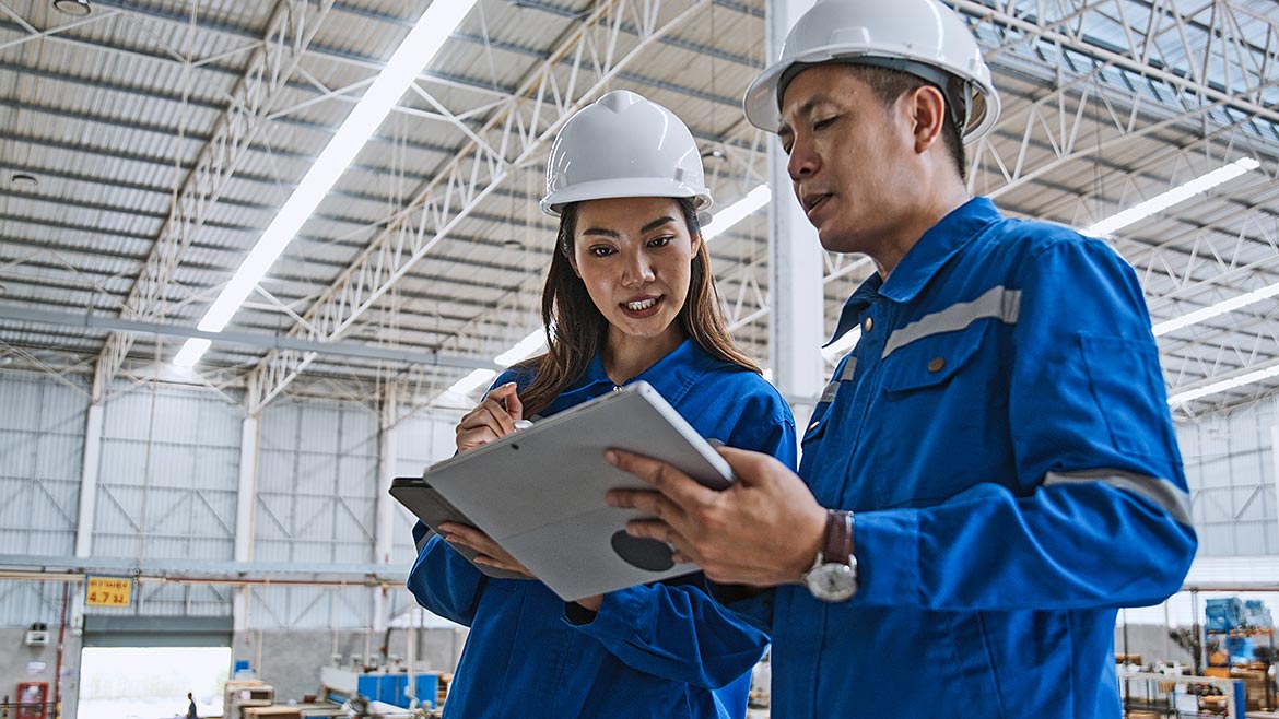 A young Asian man and woman engineer work together in a factory warehouse, using a digital tablet.