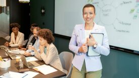 Confident young businesswoman standing and smiling at the camera in a boardroom with colleagues in the background.