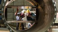 Two male workers in a metal fabrication factory inspecting a product, examining it with a flashlight.