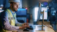 Male engineer wearing a safety vest and hard hat working on a desktop with a blurred background of the workplace.